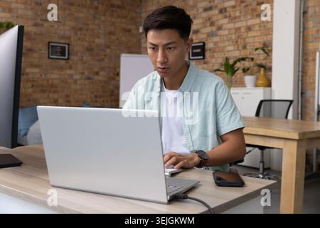 Asiatischer Mann tippt auf silbernem Laptop an einem modernen Schreibtisch im Backsteinstudio mit Monitor-Smartphone-Whiteboard. Modern, Arbeitsbereich, PR Stockfoto