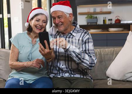 Seniorenpaar sitzt auf beigefarbener Couch im Wohnzimmer, trägt Weihnachtsmützen und sieht ein Smartphone. Familie, Urlaub, gemütlich, innen, Stockfoto