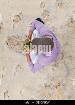 Aus der Vogelperspektive einer Person in einem lila Hemd, die an einem Sandstrand sitzt und eine grüne Kokosnuss mit Fußabdrücken im Sand hält Rio de Janeiro, Bundesstaat Rio de Janeiro Stockfoto