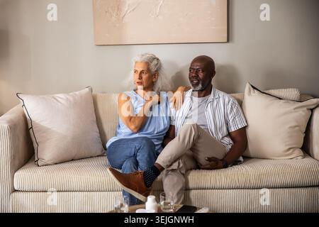 Ein vielseitiges Paar, das auf einem cremefarbenen Sofa im Wohnzimmer sitzt und mit Malerei dahinter auf sich selbst gestreckt hat. Paare, Intimität, Wärme, Komm Stockfoto