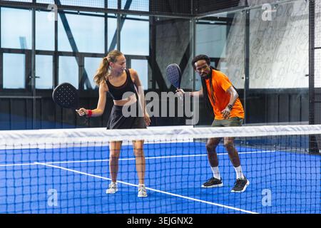 Diverse Sportpartner stehen hinter dem Tennisnetz auf dem Padelplatz in der Sporthalle und halten Schläger. Sportlich, Teamwork, Freizeit Stockfoto