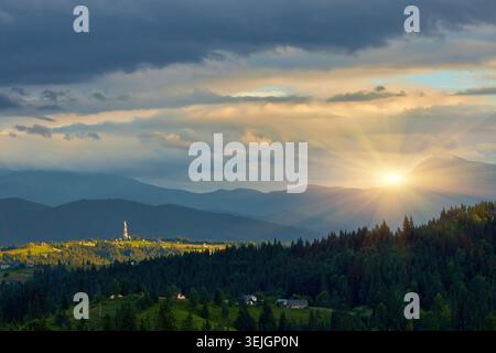 Panoramablick auf die Berglandschaft mit mehrschichtigen Graten unter einem dramatischen bewölkten Himmel. Ein sonnendurchfluteter Hügel mit einem Kommunikationsturm und Dorfständen Stockfoto
