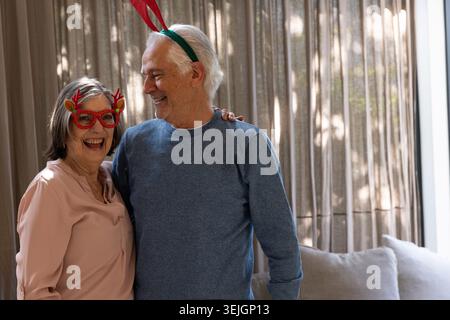 Seniorenpaar mit Rentierbrille und Geweih-Stirnband, das zu Hause mit beigen Vorhängen steht. Feier, Wärme, Begleiter Stockfoto