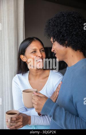 Verschiedene Paare stehen im Schlafzimmer in der Nähe des Fensters und halten strukturierte Keramikbecher und plaudern. Gemütlich, intim, Entspannung, Komfort, Stockfoto