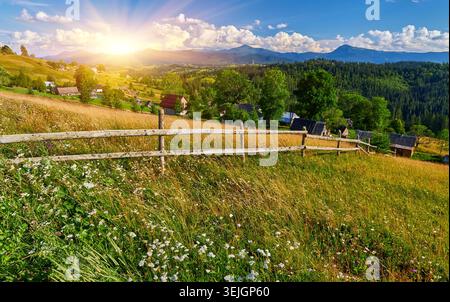 Malerischer Blick über eine Bergwiese mit Wildblumen und einem rustikalen Holzzaun. Dorfhäuser sind auf sonnigen Hängen unter bewaldeten Gipfeln und verstreut Stockfoto