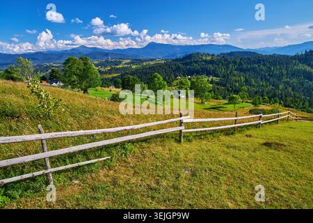 Malerischer Blick über eine Bergwiese mit Wildblumen und einem rustikalen Holzzaun. Dorfhäuser sind auf sonnigen Hängen unter bewaldeten Gipfeln und verstreut Stockfoto