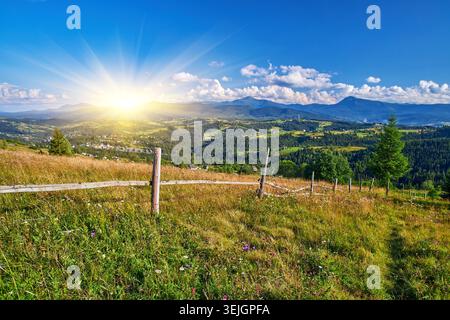 Malerischer Blick über eine Bergwiese mit Wildblumen und einem rustikalen Holzzaun. Dorfhäuser sind auf sonnigen Hängen unter bewaldeten Gipfeln und verstreut Stockfoto