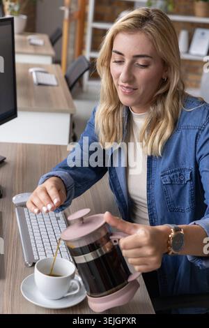 Frau im mittleren Erwachsenenalter in Denim-Hemd, die Kaffee aus rosa Französisch in die Tasse am Schreibtisch gießt. Minimalistisch, modern, Tageslicht, Stockfoto