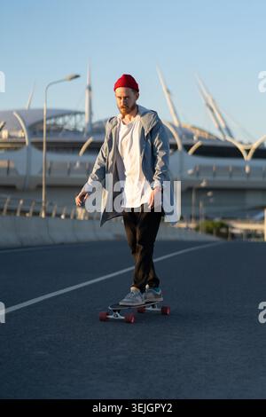 Trendiger Hipster-Mann, der draußen Schlittschuhlaufen kann. Bärtiger Millennial-Typ in Freizeitkleidung und Trendhut-Longboard im urbanen Raum bei Sonnenuntergang über dem Stadion des Stadthauses Stockfoto
