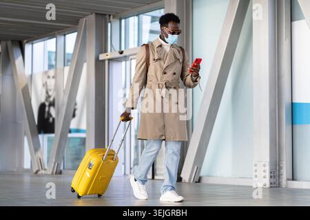 Afro-Millennial-Mann, der mit gelbem Koffer im Flughafenterminal läuft, Handy benutzt, Gesichtsmaske tragen. Reisende afrikanische ameri Stockfoto