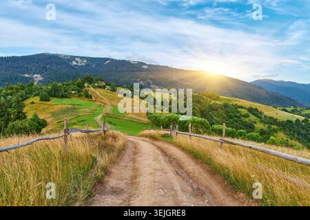 Malerische Aussicht auf einen unbefestigten Pfad, der entlang eines hölzernen Zauns auf einem hohen, grasbewachsenen Berghang verläuft. Bewaldete Hügellandschaften, die unter einem klaren blauen Himmel sichtbar sind. H Stockfoto