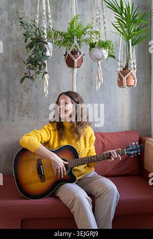 Lächelnde Freiberuflerin in gelbem Pullover, ruht auf der Couch unter Baumwoll-Makramee-Pflanzenbügel mit Zimmerpflanzen, spielt Stockfoto