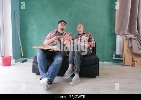 Glückliches Paar, das eine neue Wohnung renovierte, auf der Couch saß und Pizza aß, gemeinsam Pläne diskutierte und einen Schnitt streichelte Stockfoto