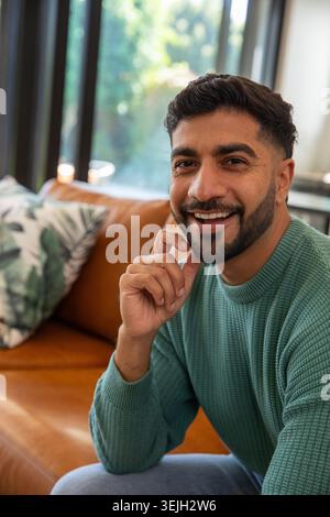Indischer Mann in 20ern sitzt zu Hause auf Ledersofa, trägt grünen Pullover, berührt das Kinn, Fenster. Gemütlich, Tageslicht, innen, grün Stockfoto