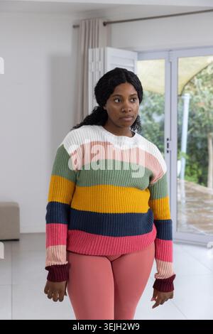 Afroamerikanerin, die zu Hause in gestreiftem Pullover und rosa Leggings neben Schiebetüren steht. Weiblich, Erwachsene, gestrickt, mehrfarbig, Muster, Mode, ca. Stockfoto
