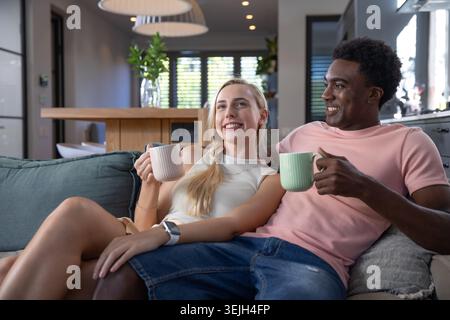 Verschiedene Paare halten Keramikbecher und lehnen sich zurück auf das türkisfarbene Sofa im modernen Wohnbereich. Gemütlichkeit, Komfort, Entspannung, Mod Stockfoto