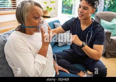 Senior Afroamerikaner Patient und asiatische Krankenschwester in Scrubs sitzend auf Sofa mit Sauerstoffmaske. Mitgefühl, Gesundheit, Wellness, Pflege, modern, KOM Stockfoto