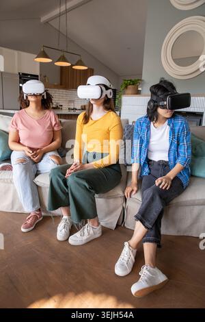 Verschiedene Freundinnen testen Virtual-Reality-Headsets, während sie im modernen Wohnzimmer auf dem Sofa sitzen. Technologie, Entspannung, Kameradschaft, modern Stockfoto