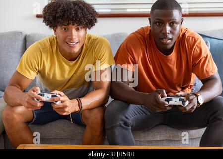 Verschiedene männliche Freunde, die sich in den Händen der Controller auf der grauen Couch zu Hause mit Jalousien, Couchtisch. Lässig, Unterhaltung, Modus Stockfoto