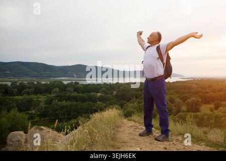 Älterer Mann mit Rucksack auf dem Gipfel des Berges mit erhobenen Händen. Ein älterer Mann, der in der Natur läuft Stockfoto