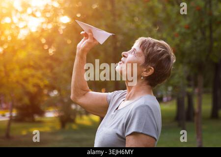 Glückliche träumliche Seniorin wirft Papierflugzeug, während sie sich draußen wünscht Stockfoto