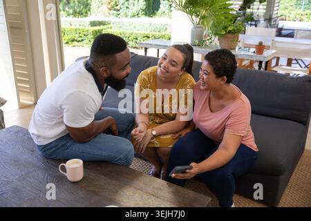 Verschiedene Freunde sitzen auf dem Sofa in der Lounge mit der Terrasse und halten ein Smartphone, während sie an einem hellrosa Becher schlürfen. Gemütlich, Freundschaft, Freizeit, Stockfoto