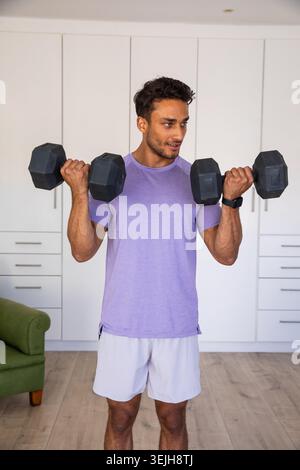 Asiatischer Mann, der schwarze Hexen-Kurzhanteln hebt und Fitness-Tracker neben grünem Sessel im Heimstudio trägt. Fitness, Kraft, Motivation Stockfoto