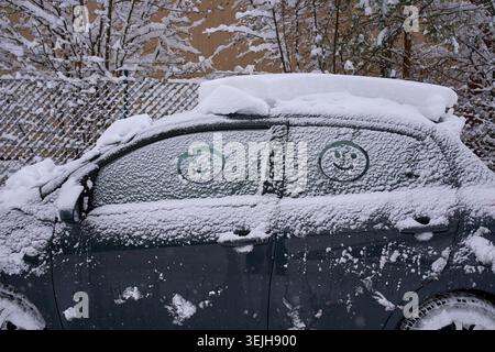 Ein schneebedecktes Auto mit lustigen Gesichtern, die im Winter auf den gefrorenen Seitenfenstern gezeichnet sind. Stockfoto
