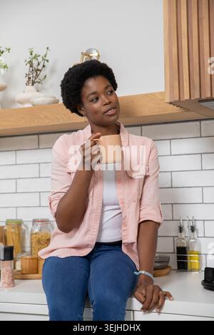 MittelErwachsene afroamerikanische Frau, die auf der Arbeitsplatte in der Küche sitzt und einen pfirsichfarbenen Keramikbecher hält. Modern, minimalistisch, gemütlich, Stockfoto