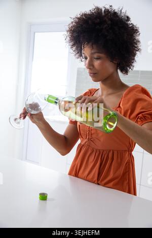 Afroamerikanerin in orangefarbenem Kleid, die Wein aus der grünen Flasche in das Glas in der Hausküche gießt. Eleganz, Freizeit, rustikal, Lifestyle, Geselligkeit, Stockfoto