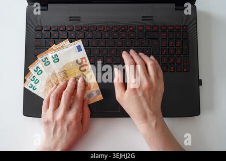 Eine Hochwinkelansicht der Hände, die fünfzig Euro-Scheine halten, während sie auf einer schwarzen Laptop-Tastatur tippen. Stockfoto
