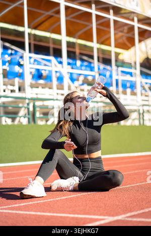 Durstige Frau, die Wasser aus einer Plastikflasche trinkt, ruht sich nach dem Joggen auf einem Gummistadion des Laufbands an sonnigen Sommertagen aus Stockfoto