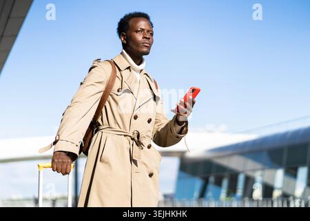 Afro-amerikanischer Millennial Traveler Mann mit gelbem Koffer steht im Flughafenterminal, hält Telefon, ruft an und sucht nach einem Stockfoto