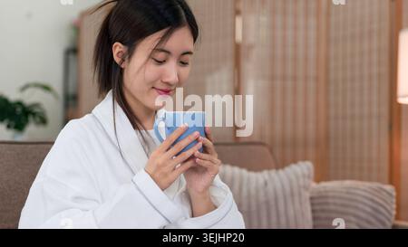 Asiatische Frau, die weißen Bademantel trägt, hält blaue Tasse, die heißes Getränk trinkt und sich morgens im Wohnzimmer zu Hause entspannt Stockfoto