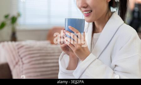 Asiatische Frau im Bademantel trinkt Kaffee, während sie sich auf dem Sofa während der morgendlichen Routine zu Hause entspannt und friedliche Momente genießt Stockfoto