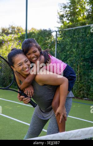 Asiatische Mutter und Tochter in Sportbekleidung, die Huckepack auf dem Tennisplatz reiten, während sie Schläger halten. Familie, Aktivität, Freizeit, f Stockfoto