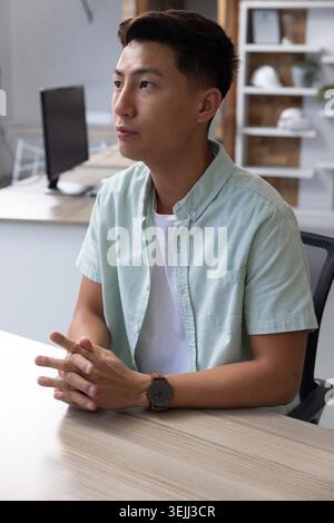 Asiatischer Mann mit mintgrünem Hemd, der am Schreibtisch im Büro sitzt und mit Schutzhüten in die Regale blickt. Professionell, Arbeitsplatz, Minimalismus, Innenraum, Co Stockfoto