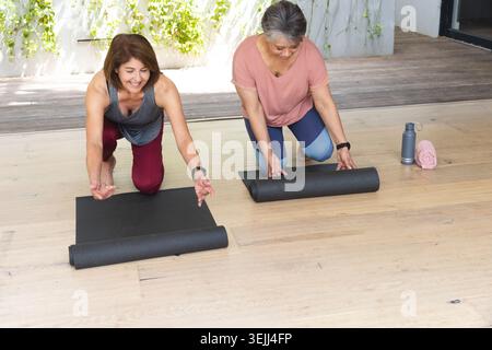 Hispanische und ältere asiatische Frauen, die schwarze Yogamatten auf der Terrasse mit Wasserflasche und rosafarbenem Handtuch ausrollen. Wellness, Fitness, Begleitung Stockfoto