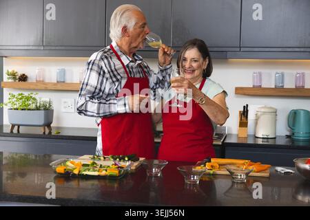 Ein älteres Paar trägt rote Schürzen und schneidet Gemüse auf einer Steinkücheninsel mit Weingläsern. Küche, Kochen, Kulinarik, ru Stockfoto