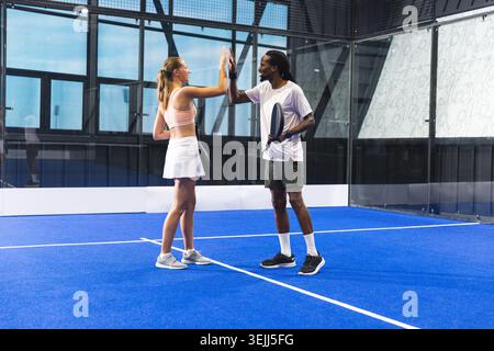 Verschiedene Teamkameraden stehen sich in der Nähe des Netzes auf einem blauen Hallenpadelplatz gegenüber, Schläger in der Hand. Sport, Teamwork, Wettkämpfe Stockfoto