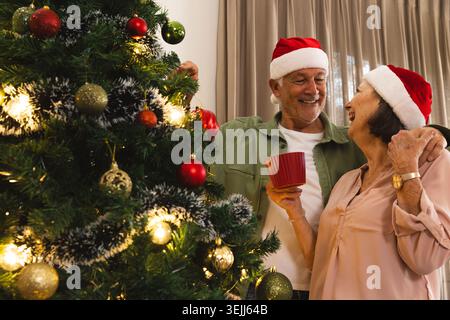 Ältere Ehepaare mit Weihnachtsmützen schmücken zu Hause einen Weihnachtsbaum, halten rote Tassen in den Händen, Platz für Text. Festiv Stockfoto