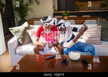 Verschiedene männliche Freunde sitzen zuhause auf dem Sofa, tragen VR-Headsets, halten Controller in der Hand und mit Popcorn n Stockfoto