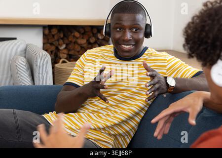Verschiedene Freunde unterhalten sich über Over-Ear-Kopfhörer, während sie auf einem blauen Stoffsofa im Wohnzimmer sitzen. Gemütlichkeit, Unterhaltung, Stockfoto