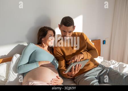 Verschiedene Paare sitzen aufrecht auf dem Bett im Schlafzimmer, betrachten zusammen eine Tablette und berühren sich gegenseitig die Mägen. Roman Stockfoto
