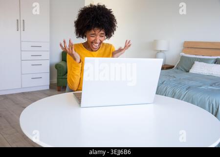 Afroamerikanische Frau in senfgelber Oberseite feiert mit Laptop am Tisch im Schlafzimmer. Freude, Leistung, Heimbüro, gemütlich, Stockfoto