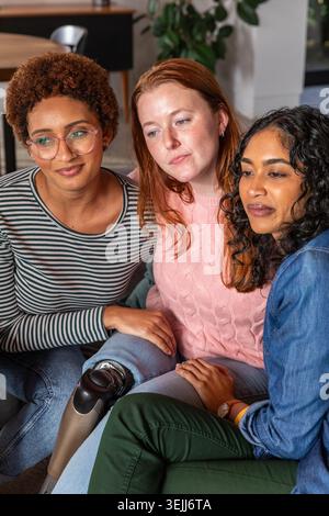 Verschiedene Freundinnen sitzen zu Hause auf dem Sofa, tragen eine Brille und zeigen ihre Beinprothesen. Kameradschaft, Sicherheit, also Stockfoto