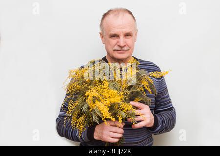 Der ältere Mann mit einem Blumenstrauß aus Frühlingsblumen einer Mimosa. Überraschung für Liebling. Geburtstag. Stockfoto