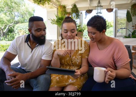 Verschiedene Freunde sitzen auf einem Sofa unter einer Pergola, zeigen auf eine Tablette und halten eine Kaffeetasse in der Hand. lou im Freien Stockfoto