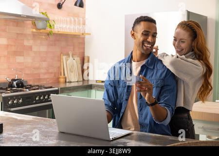 Verschiedene Paare teilen sich einen Moment zu Hause auf der Kücheninsel, während sie einen Laptop und ein Smartphone in der Nähe des Gasherds verwenden. Familie, Konv Stockfoto