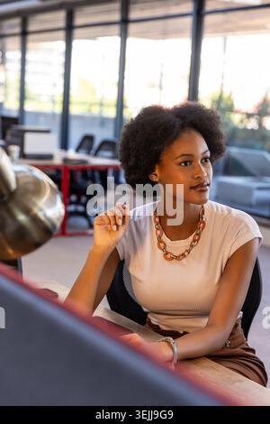 Afroamerikanische Frau, die am Schreibtisch sitzt und Bleistift hält und auf den Monitor in der Nähe einer Metalllampe blickt. Konzentration, Kreativität, in Stockfoto